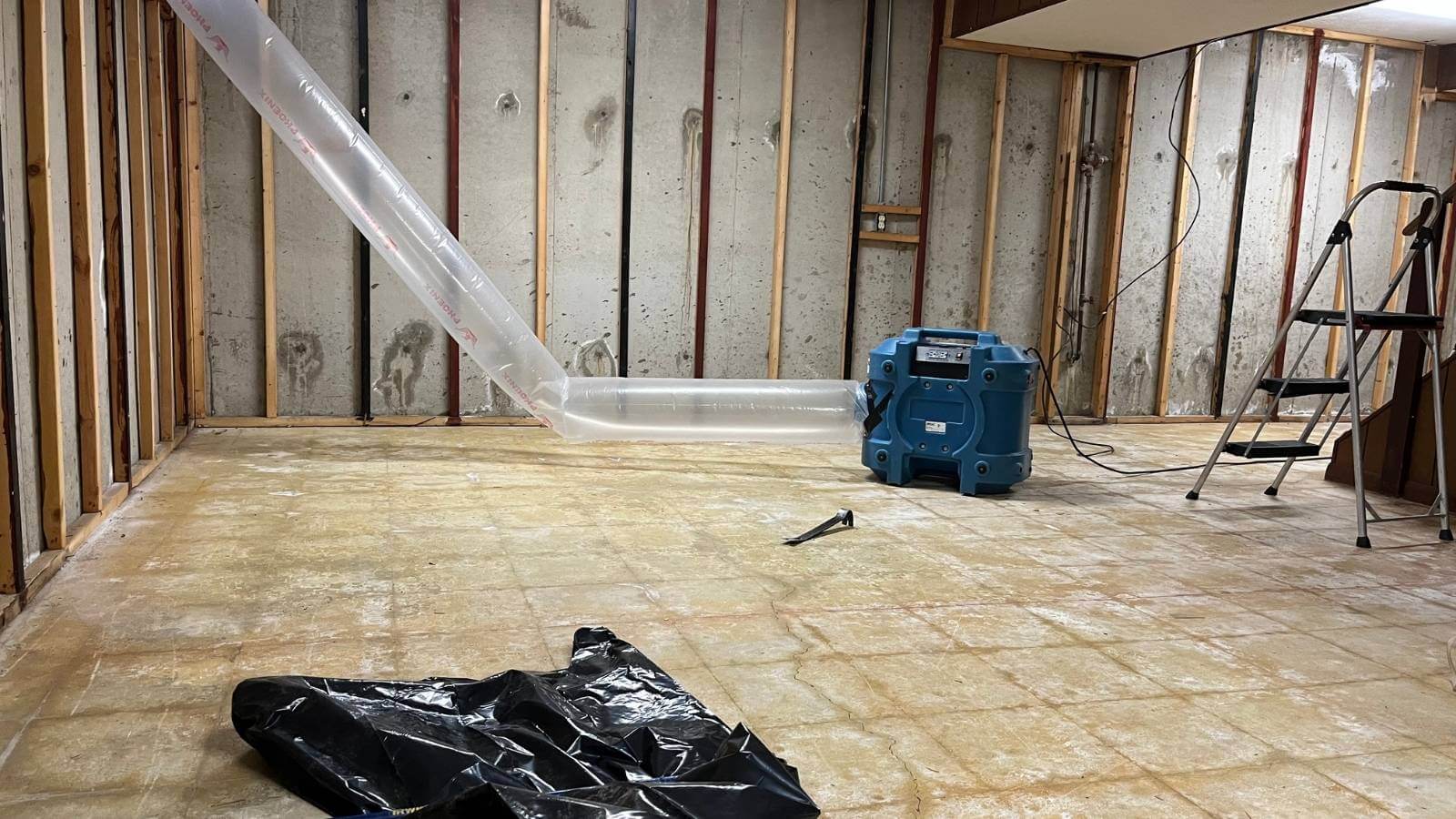 A basement undergoing restoration, featuring exposed walls, a blue dehumidifier, and a clear plastic tube for moisture removal.