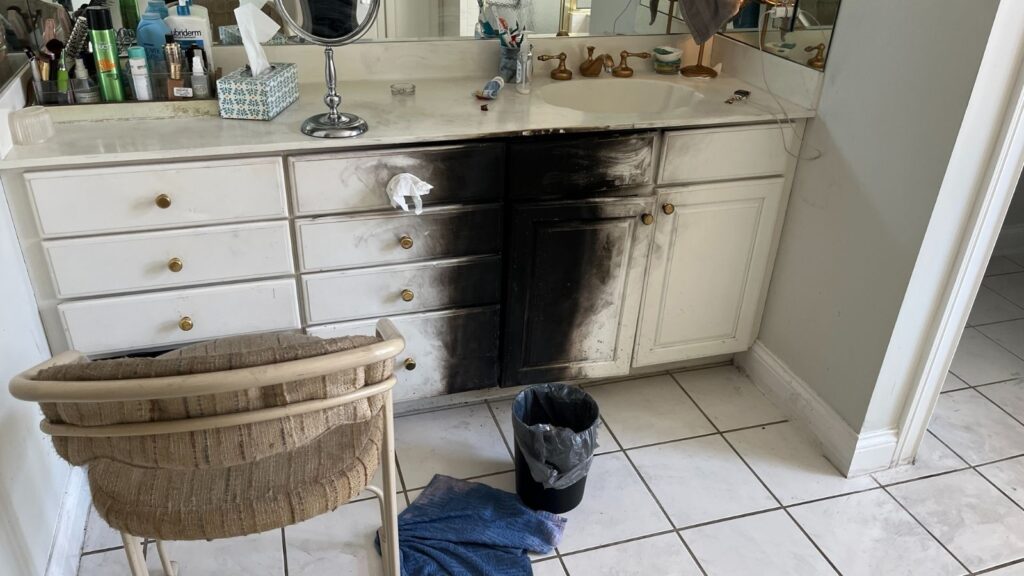 A bathroom vanity with one side charred black from fire damage, a wicker chair, garbage can, and cleaning cloth on the tiled floor.