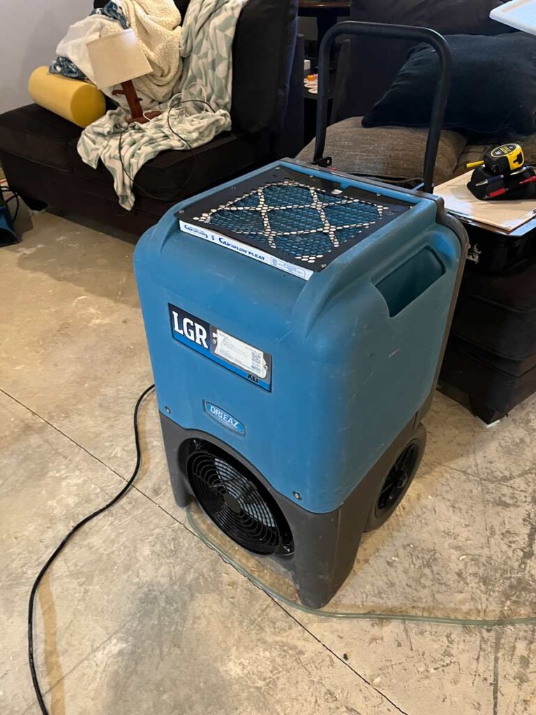 A blue dehumidifier sits on a concrete floor beside a sofa, with a blanket and lamp nearby, in a cluttered living space.