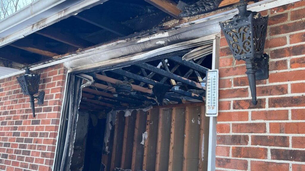 Exterior view of a brick home with fire damage, showing charred beams and lanterns, and a damaged wall inside.