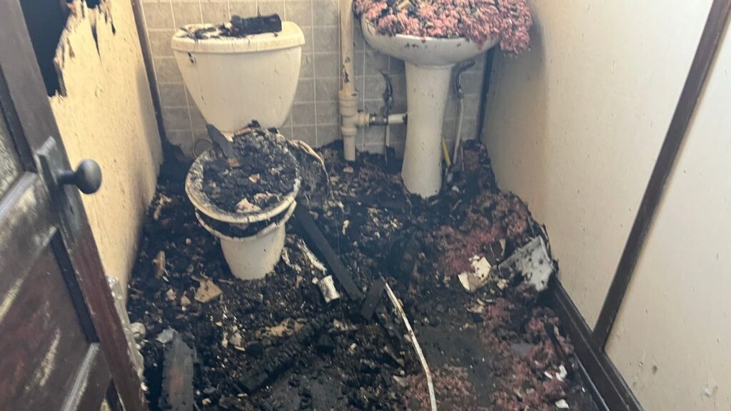 A damaged bathroom with charred fixtures and debris, showing signs of fire damage on walls and floors.