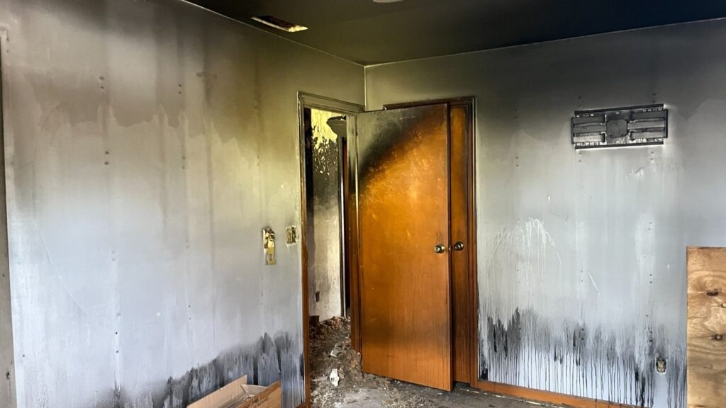 A room with charred walls and a blackened door, indicating significant fire damage, with soot stains and debris on the floor.