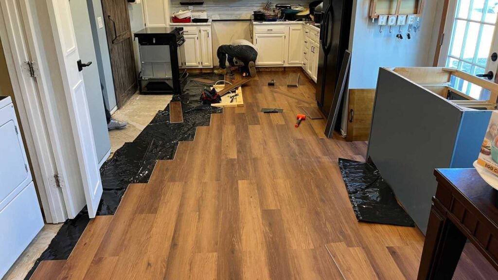 A person installs new wooden flooring in a kitchen, surrounded by tools and plastic sheeting, with cabinets and appliances nearby.