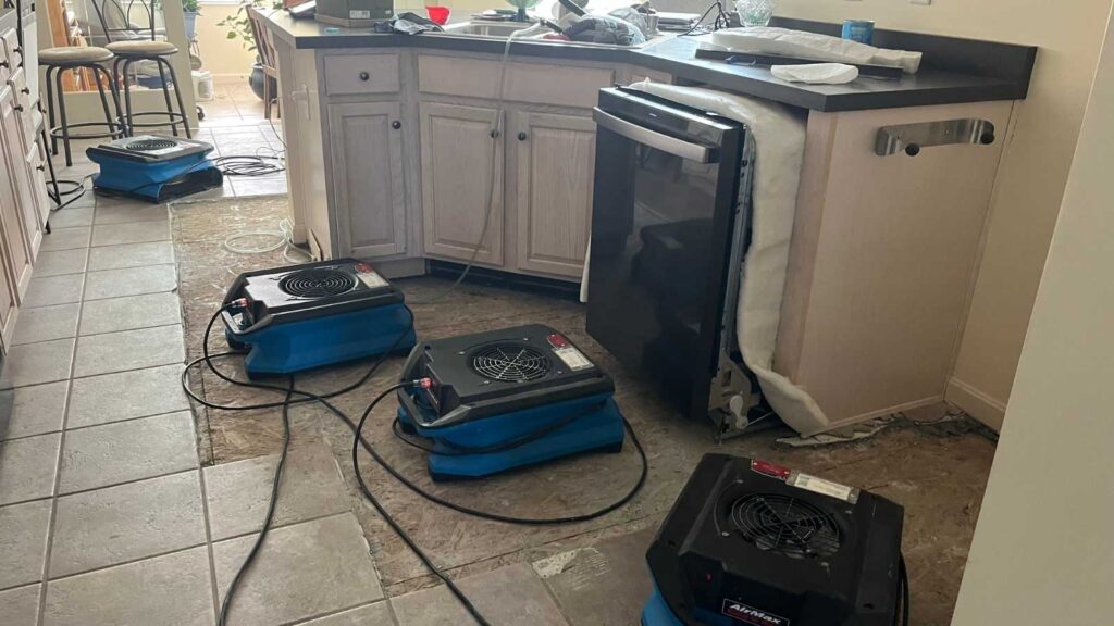 Three air movers are placed on a kitchen floor amid renovation, with exposed flooring and a refrigerator being serviced.