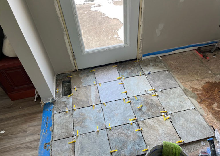 Newly laid slate tiles on a floor near a door, with spacers in place for grout. Adjacent area shows unfinished flooring and tools.