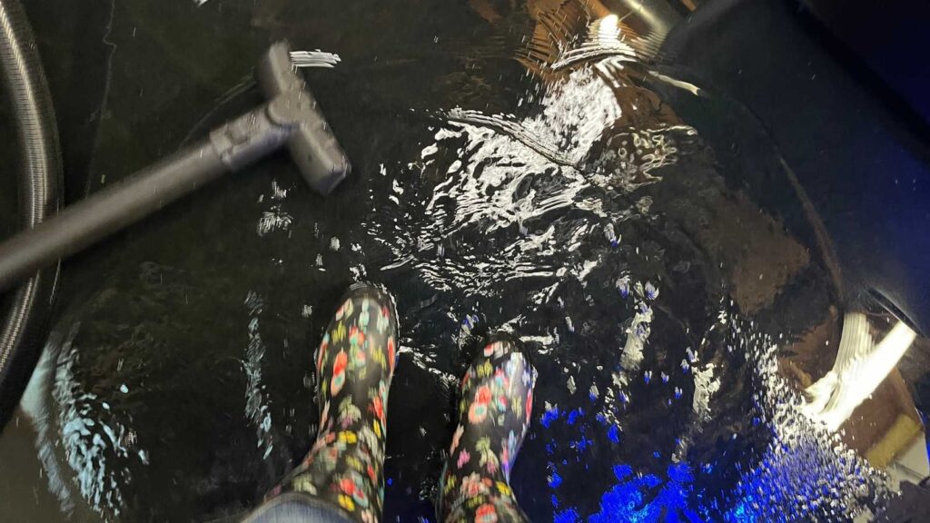 Colorful rain boots stand in a pool of water, with a vacuum nearby, reflecting light on a glossy surface.