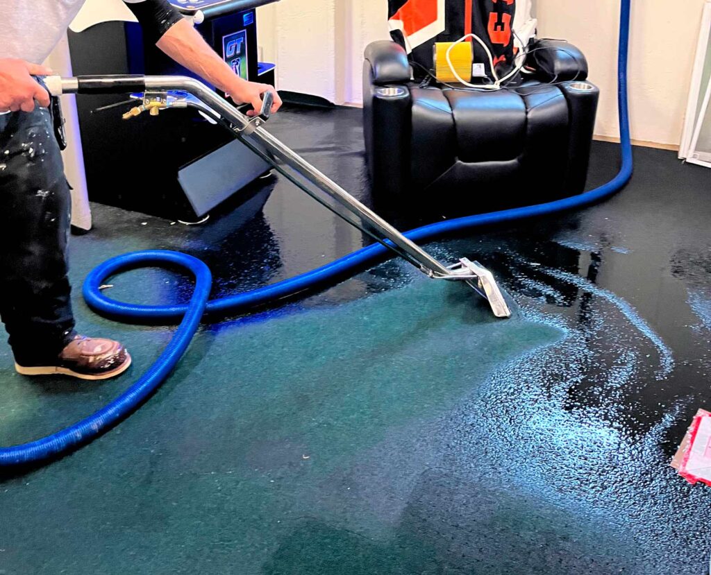 A person cleans a dark carpet with a carpet cleaner, while a blue hose sprawls across the floor, beside a black reclining chair.