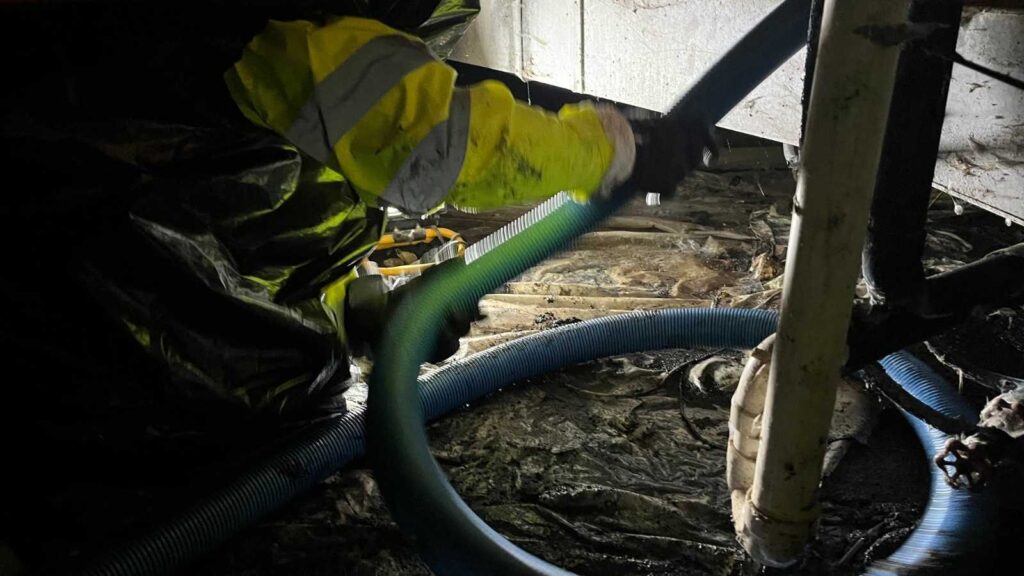 A worker in a reflective jacket uses a green and blue hose, cleaning a dark, muddy area beneath a structure.