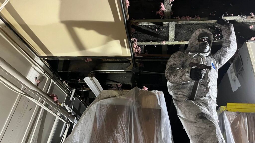 A worker in a protective suit and mask holds a tool while inspecting a damaged ceiling with insulation debris visible.