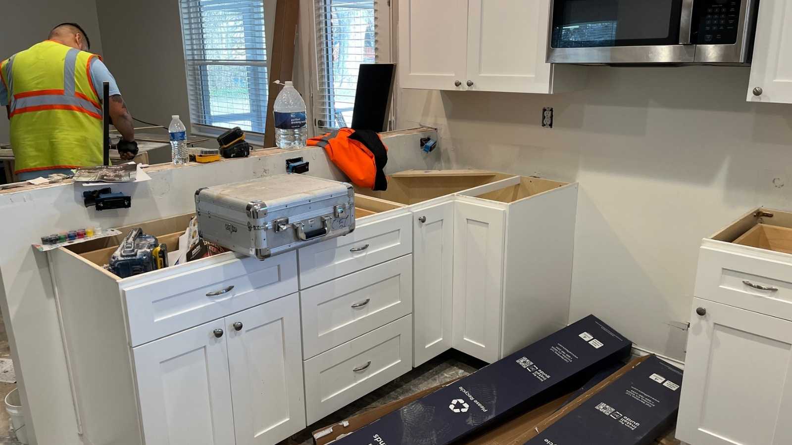 A worker in a safety vest is assembling white kitchen cabinets in a partially renovated kitchen with tools and materials scattered around.