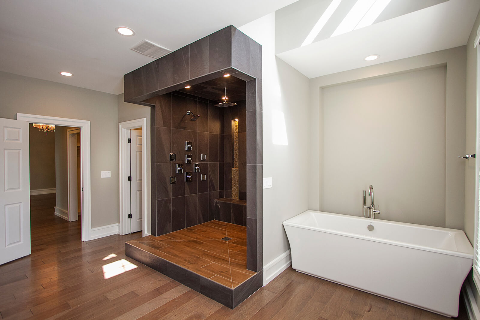 Spacious, modern bathroom with dark hardwood floors and light grey walls. It features an open, walk-in shower with dark brown large format tiles, multiple showerheads, and a built-in bench, elevated on a step. A contemporary white freestanding bathtub with a floor-mounted faucet is positioned next to the shower structure. An open doorway leads to another room.