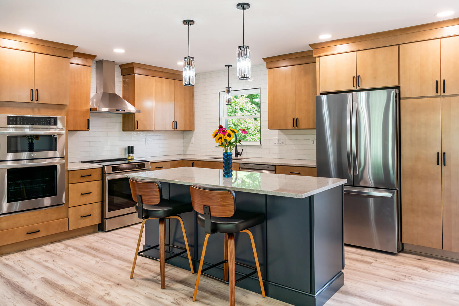 Bright, modern kitchen with light wood-toned cabinets and stainless steel appliances, including an oven, microwave, and refrigerator. It features a large kitchen island with a dark blue base, light gray countertop, and two wooden and black leather bar stools. The backsplash is white subway tile, and two modern pendant lights hang above the island. A window above the sink looks out onto green foliage.